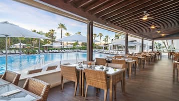 A poolside restaurant with wicker furniture, large umbrellas, and a view of the pool and palm trees.
