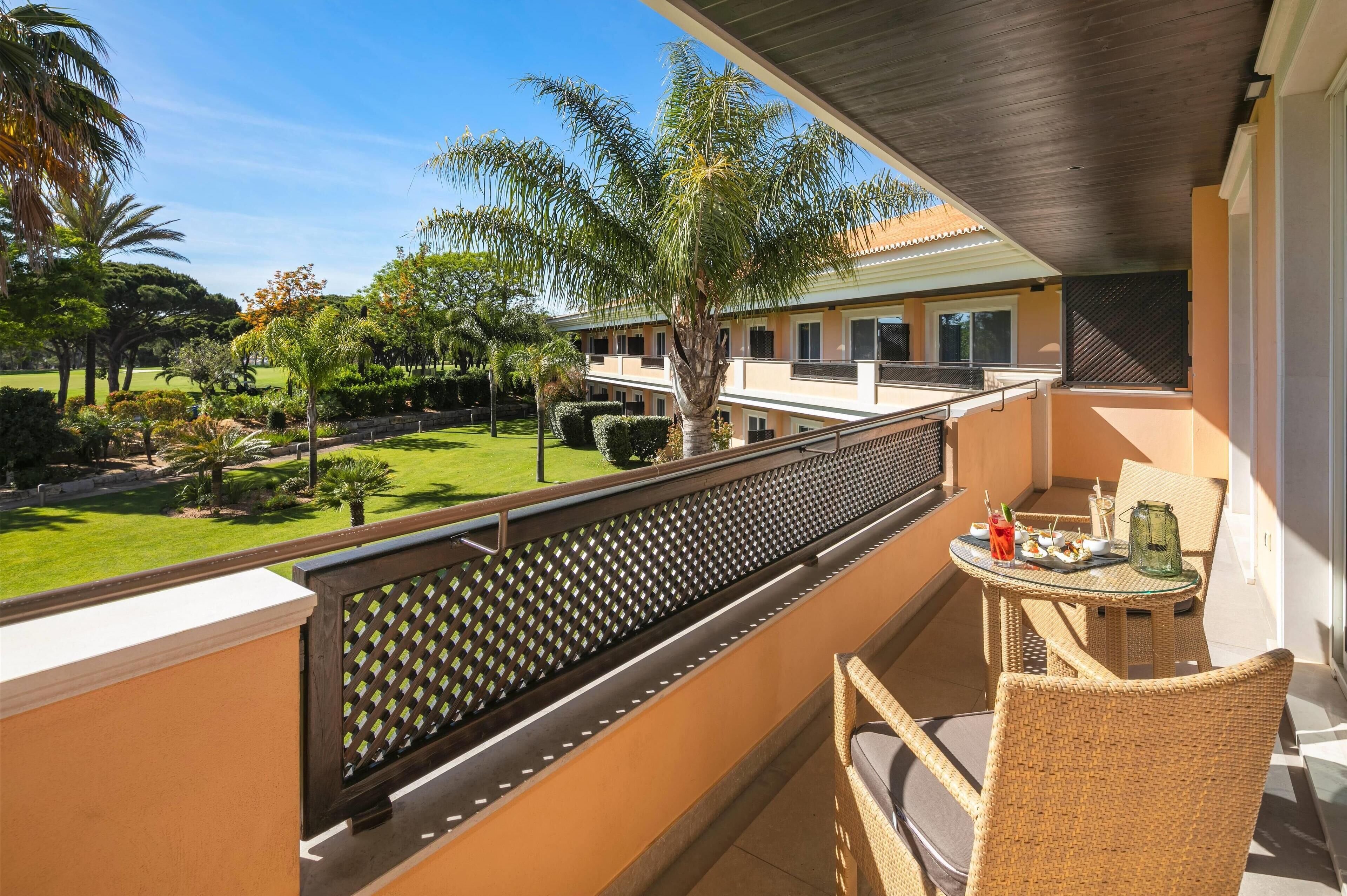 A balcony with wicker furniture, a table with a tray, and a view of a well-maintained garden with palm trees and a building in the background.