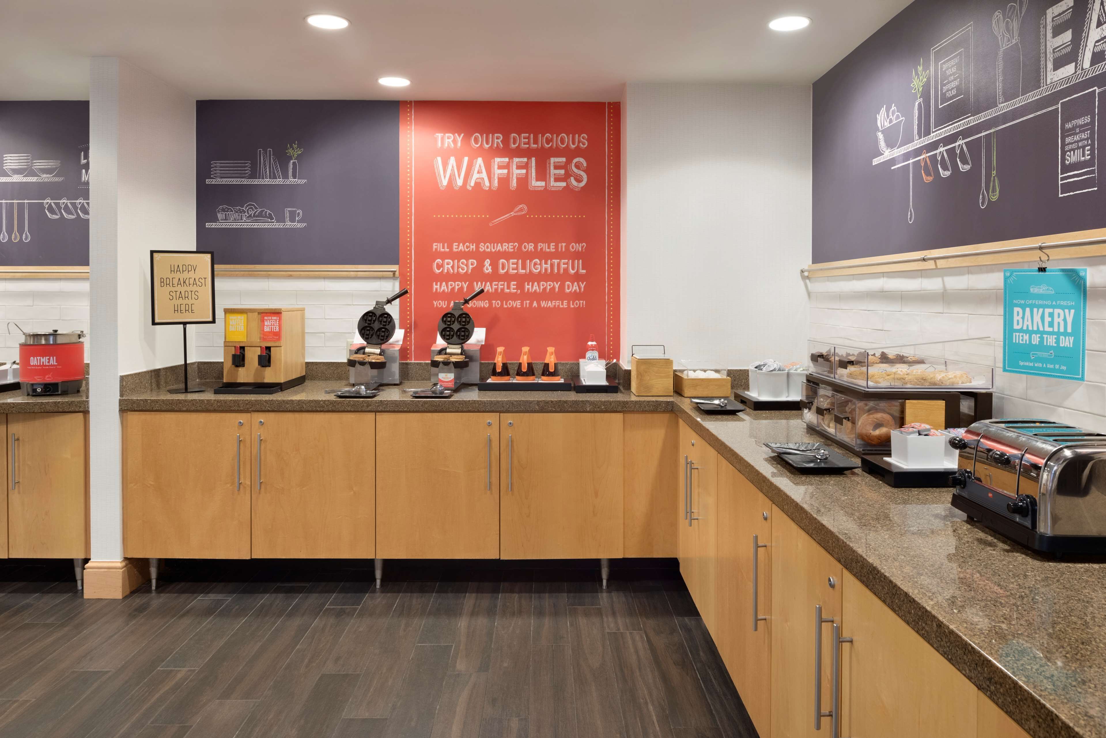 Un espace réservé au petit-déjeuner avec des postes de préparation de gaufres, un panneau publicitaire faisant la promotion des gaufres et un présentoir présentant la pâtisserie du jour.
