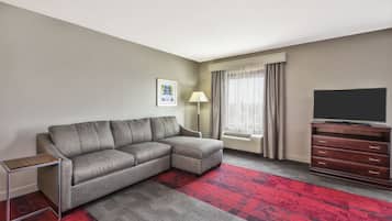A modern hotel room with a grey sectional sofa, a small side table, a wooden dresser, and a flat-screen TV.