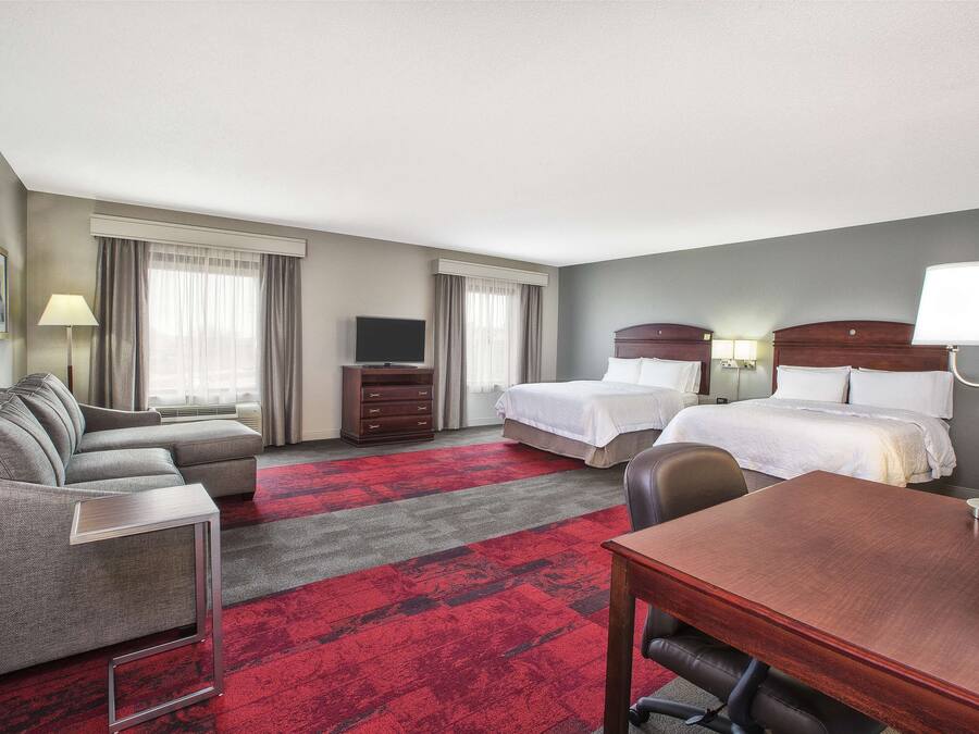 A hotel room with a sofa, two beds, a TV, a desk, and a red carpet.
