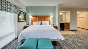A hotel room with a large bed, two bedside lamps, a wooden headboard, a small bedside table, a bench, a large window, and a view of the bathroom and kitchen. - Hampton Inn Wilmington - University Area/Smith Creek Station (Wilmington)