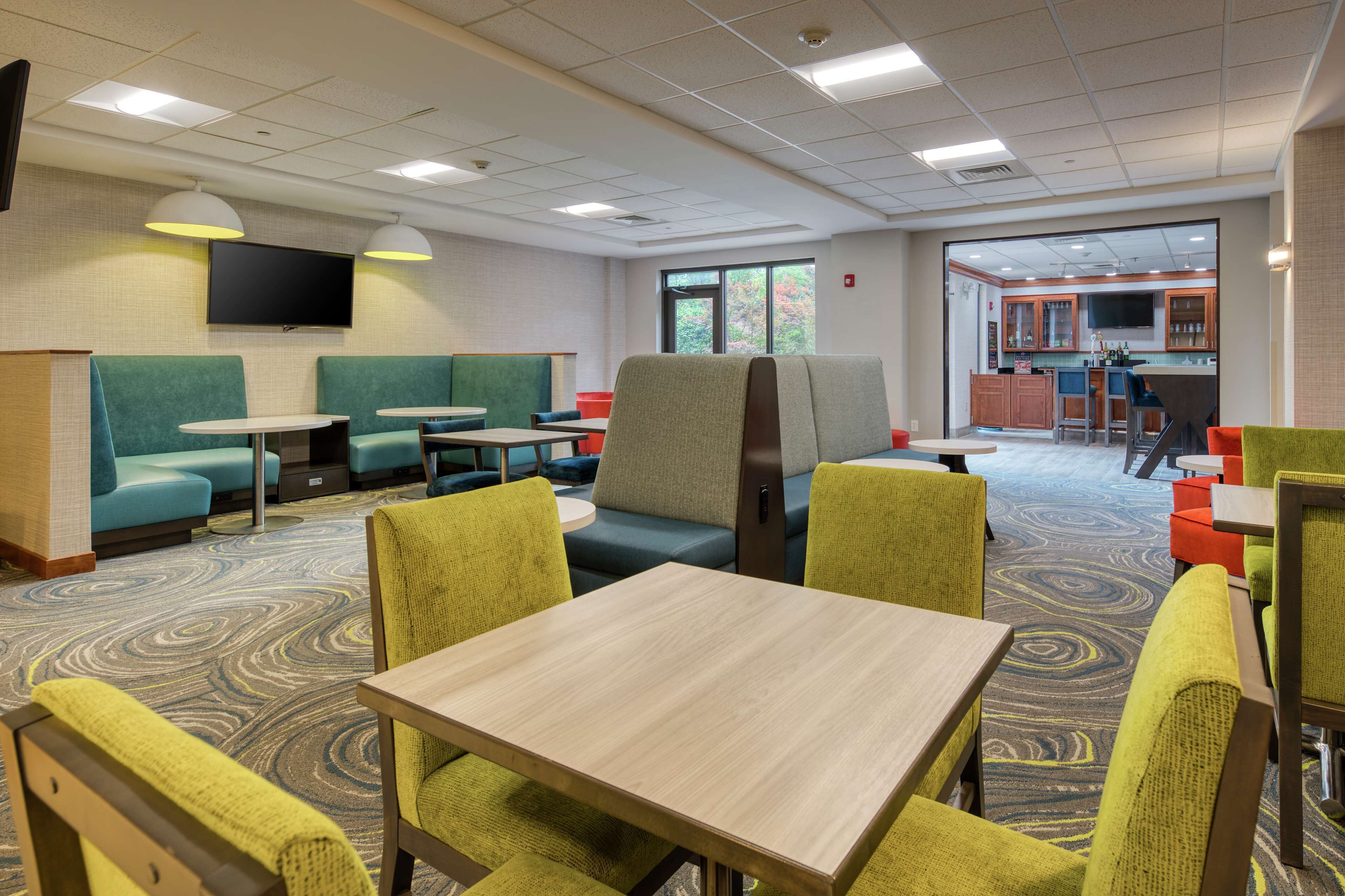 A hotel room with green seating, a flat-screen TV, a kitchen area, and a patterned carpet.