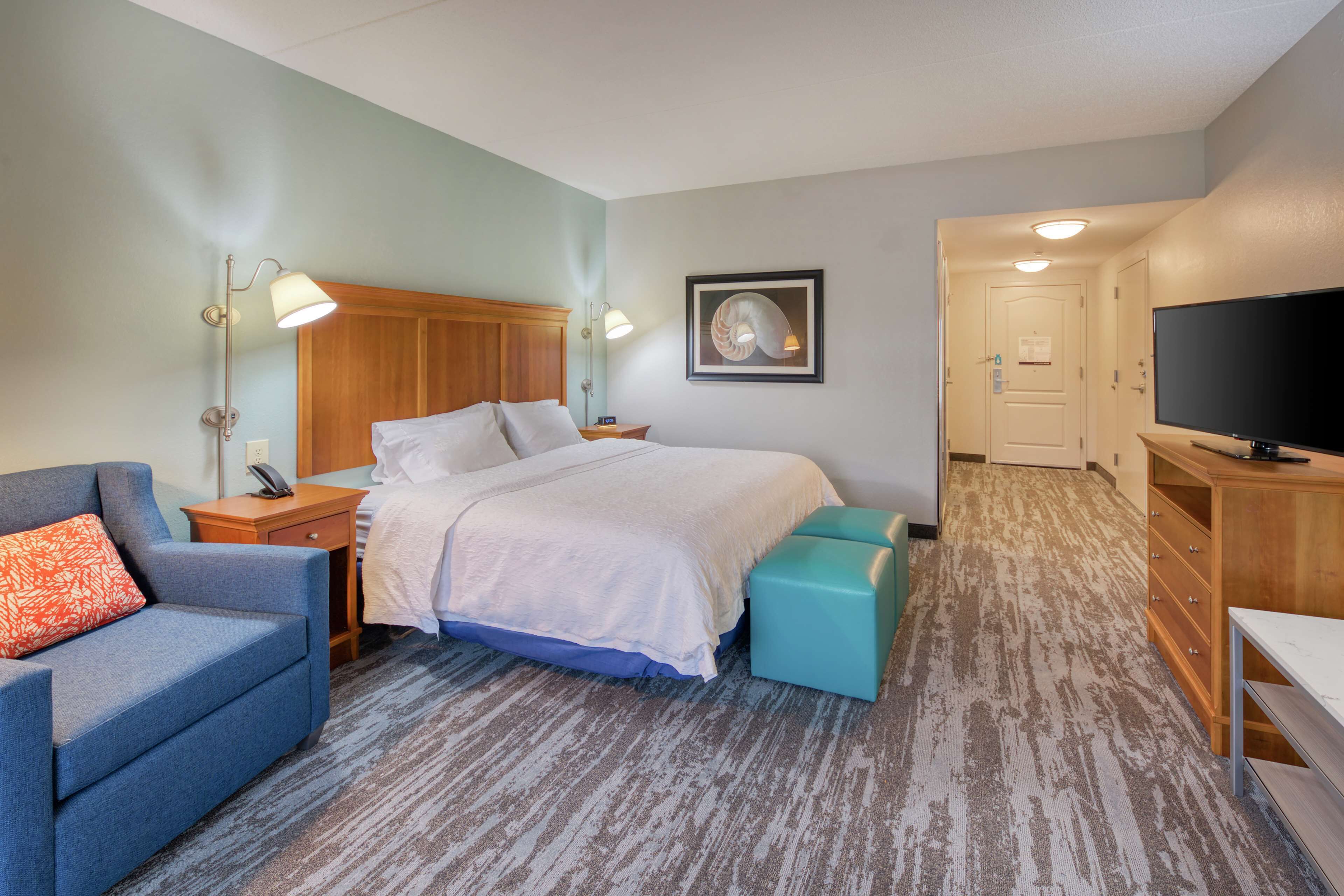 Una habitación de hotel con una cama, un sillón azul, un reposapiés verde azulado, una mesita de noche de madera, una lámpara, un cuadro en la pared, un televisor y una puerta que da a otra habitación.