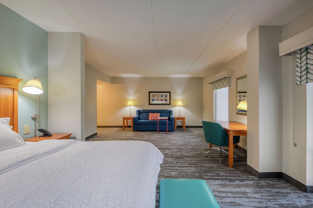Ein Hotelzimmer mit einem großen Bett, einem Schreibtisch, einem Stuhl und einer Couch. An beiden Seiten der Couch stehen jeweils zwei Lampen.