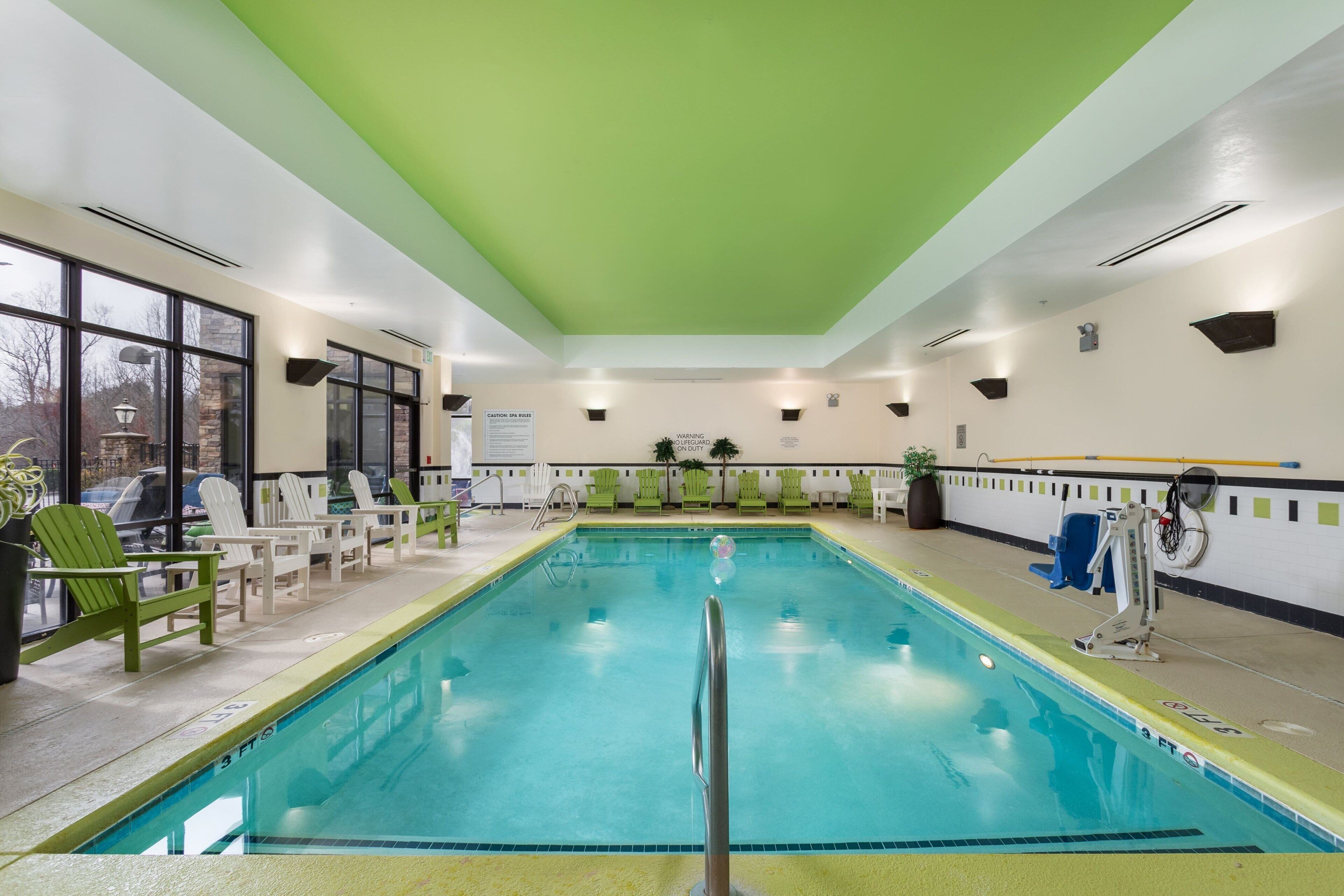 Una piscina interior con agua cristalina, rodeada de sillones de descanso verdes y blancos, y un techo verde.