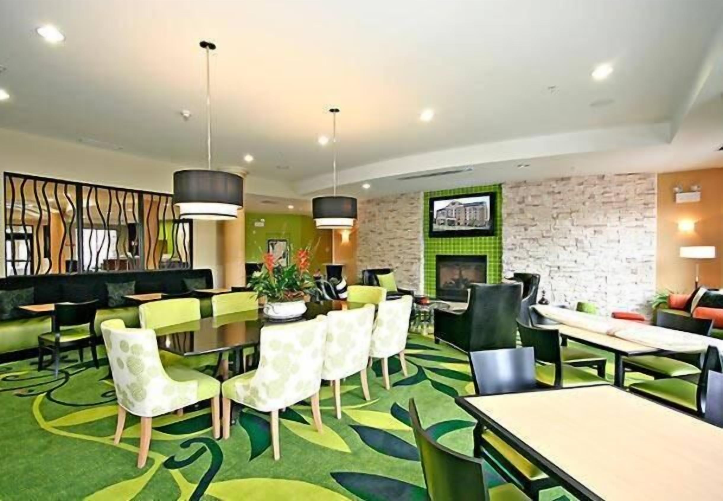 A hotel lobby with a green carpet, multiple dining tables, chairs, and a fireplace.