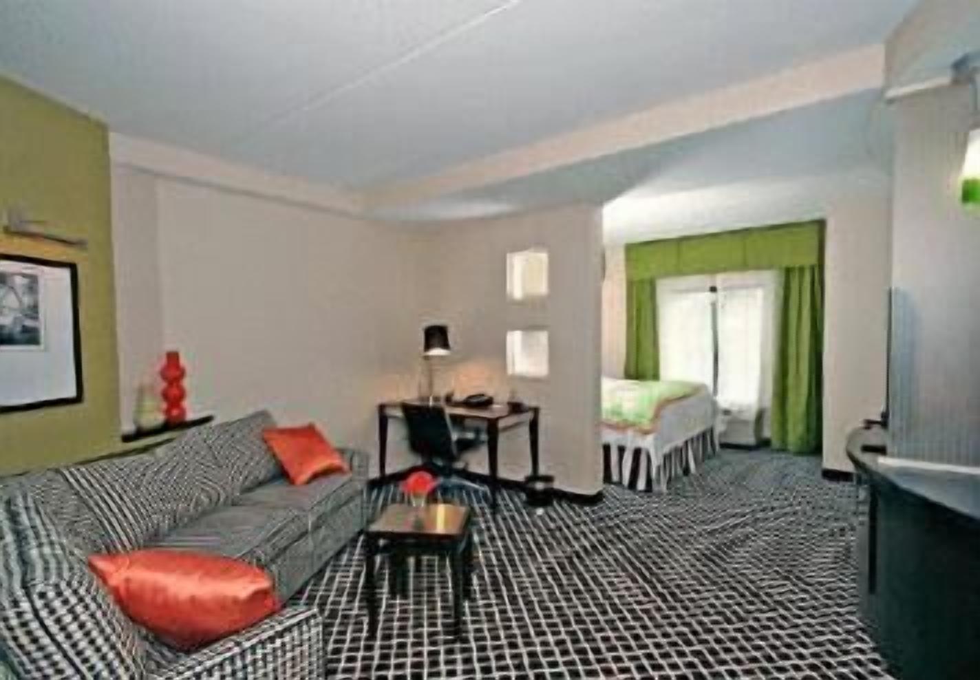 Una habitación de hotel con piso a cuadros, un sofá con cojines rojos, una mesita con una lámpara y una cama con cortinas verdes.