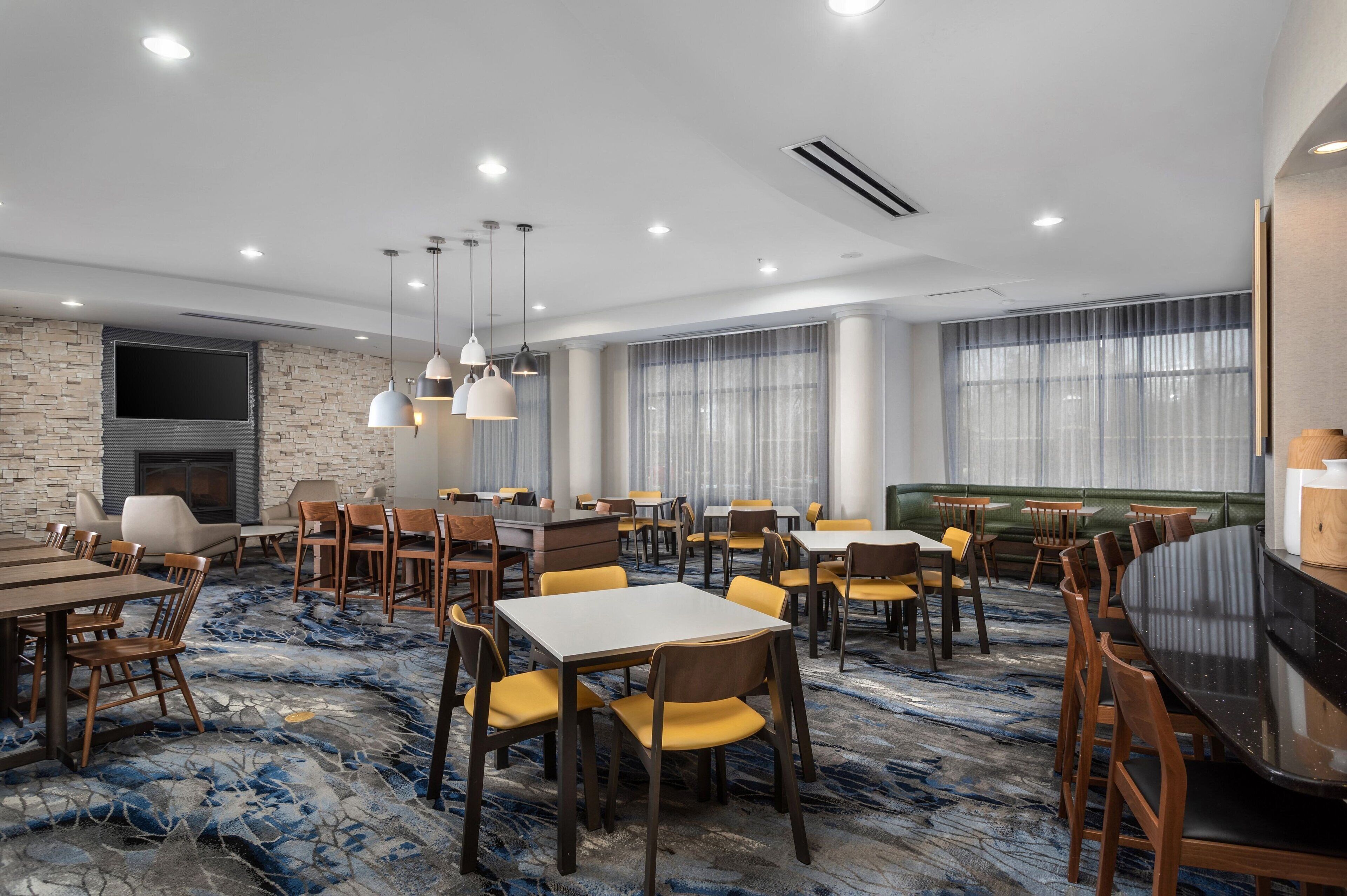 A hotel dining area with multiple tables and chairs, a fireplace, and large windows with curtains.