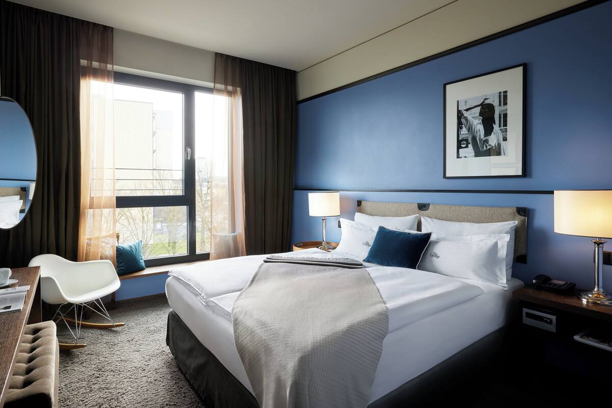 Habitación de hotel con una cama grande, dos mesitas de noche, un escritorio con una silla, una ventana con cortinas e imagen enmarcada en la pared.