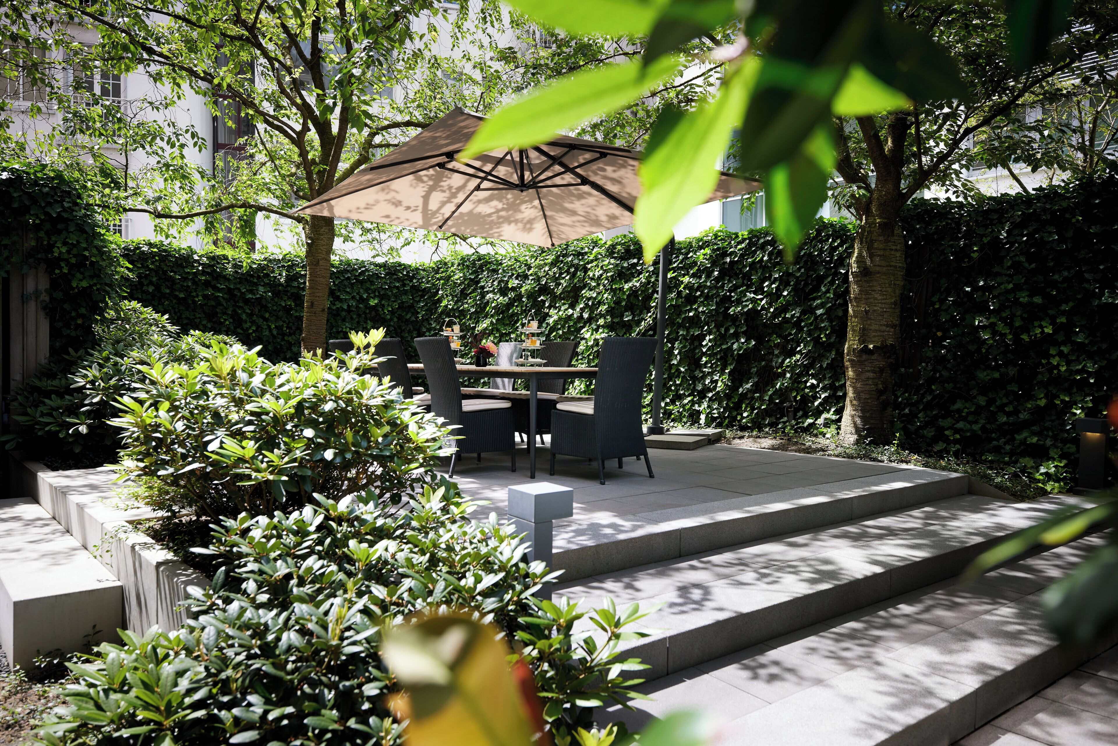 Eine Außenpatio mit Tisch und Stühlen unter einem großen Sonnenschirm, umgeben von üppiger Bepflanzung und einem Kiesweg.