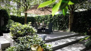 Eine Außenpatio mit Tisch und Stühlen unter einem großen Sonnenschirm, umgeben von üppiger Bepflanzung und einem Kiesweg.