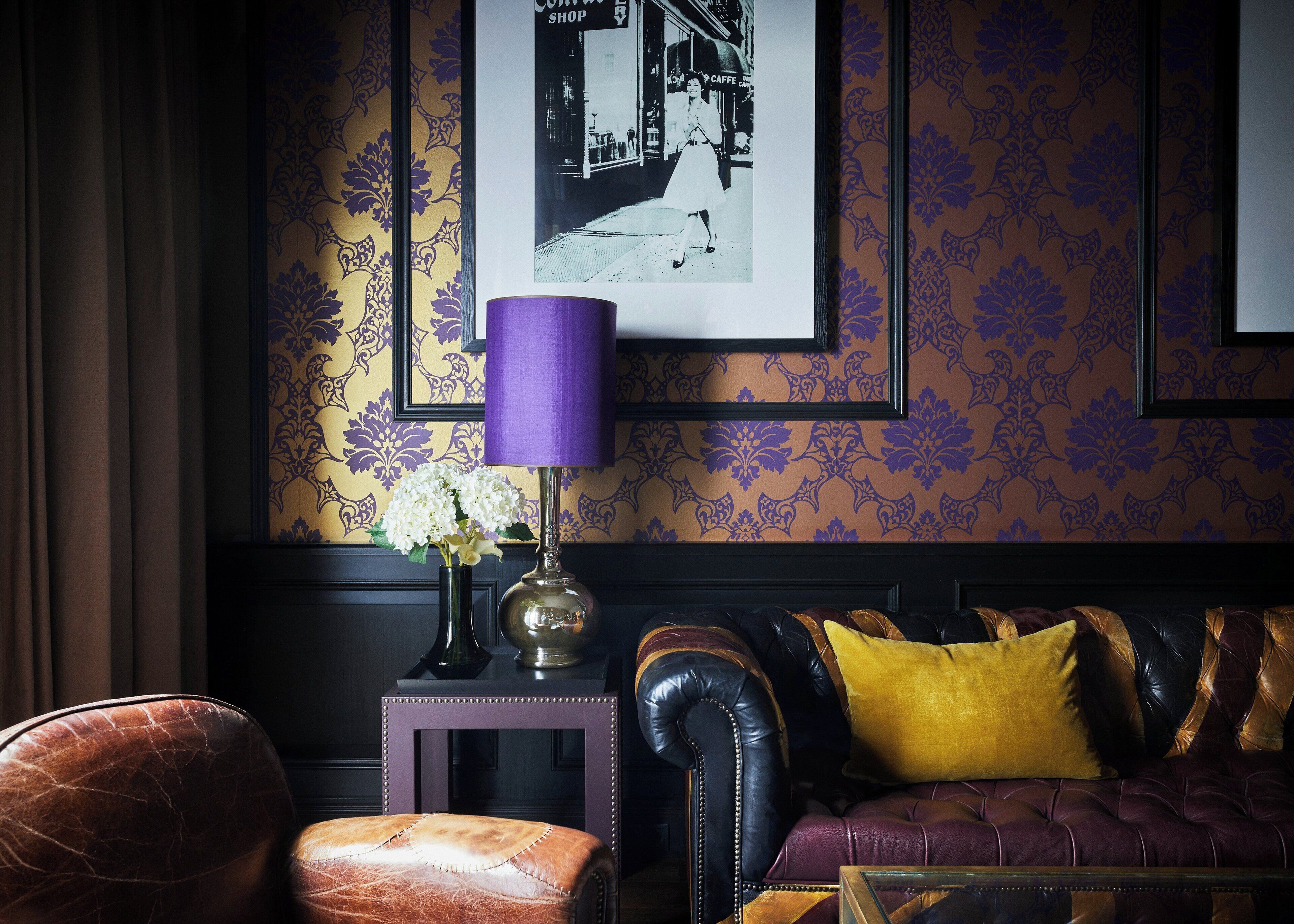 Una habitación con papel tapiz estampado, un sofá de cuero negro con cojines amarillos, una mesita auxiliar con una lámpara y una fotografía enmarcada en la pared.