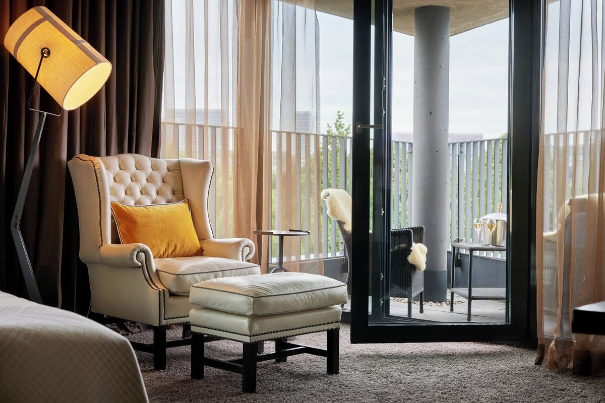 Una habitación de hotel con un ventanal grande, un sillón cómodo, una mesita y una lámpara.