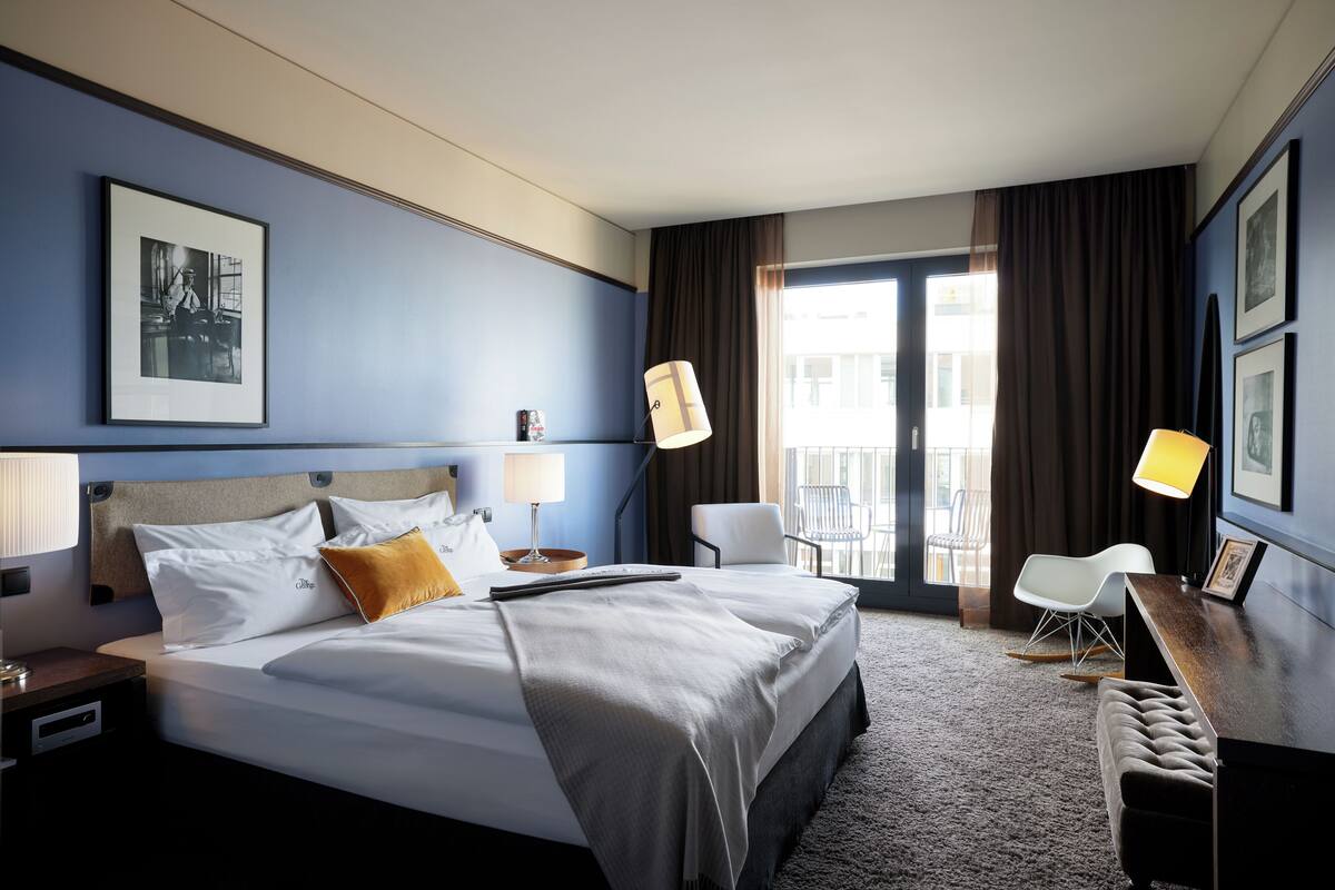 Habitación de hotel con una cama grande, dos mesitas de noche, un escritorio con silla, un balcón con vistas y un cuadro en la pared.