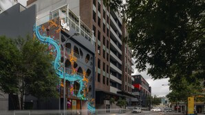 Un edificio de varios pisos con un mural colorido en la fachada, una acera y una calle con automóviles.