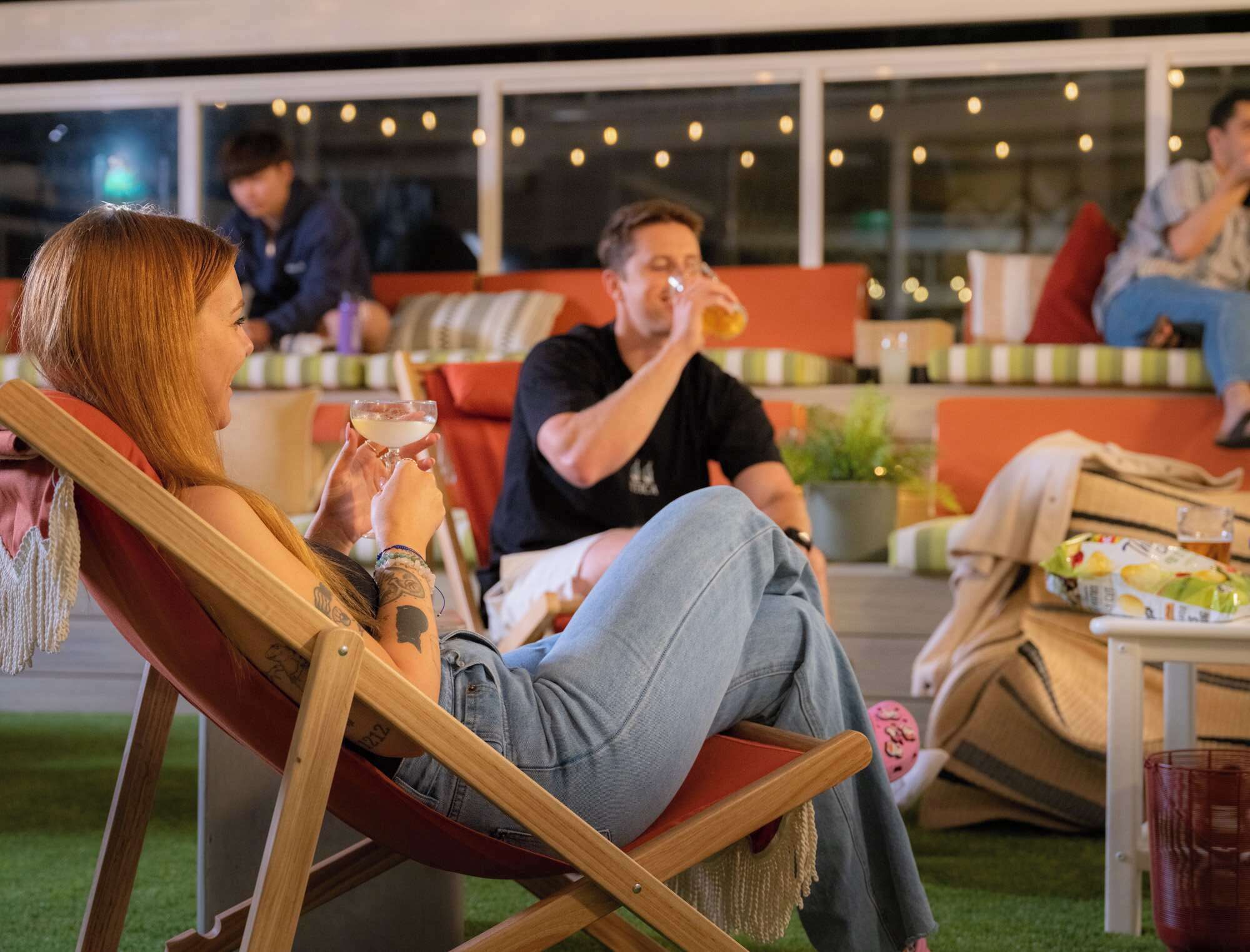 A person sitting on a wooden chair outdoors with a drink in hand, another person in the background, and a cozy seating area with cushions and a table.