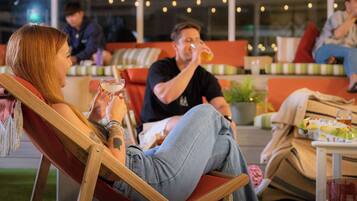 A person sitting on a wooden chair outdoors with a drink in hand, another person in the background, and a cozy seating area with cushions and a table.
