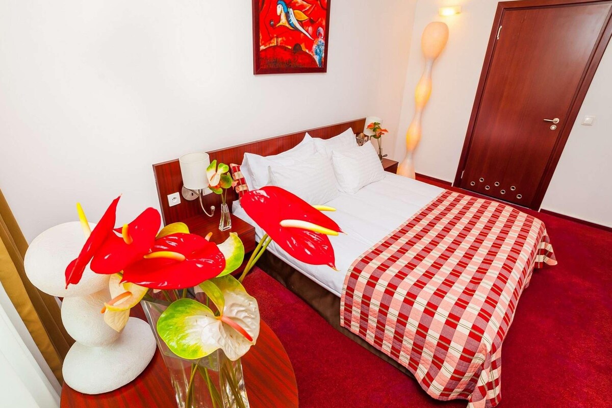 A hotel room with a bed, a red checkered bedspread, a vase with red flowers, a painting on the wall, and a wooden door.
