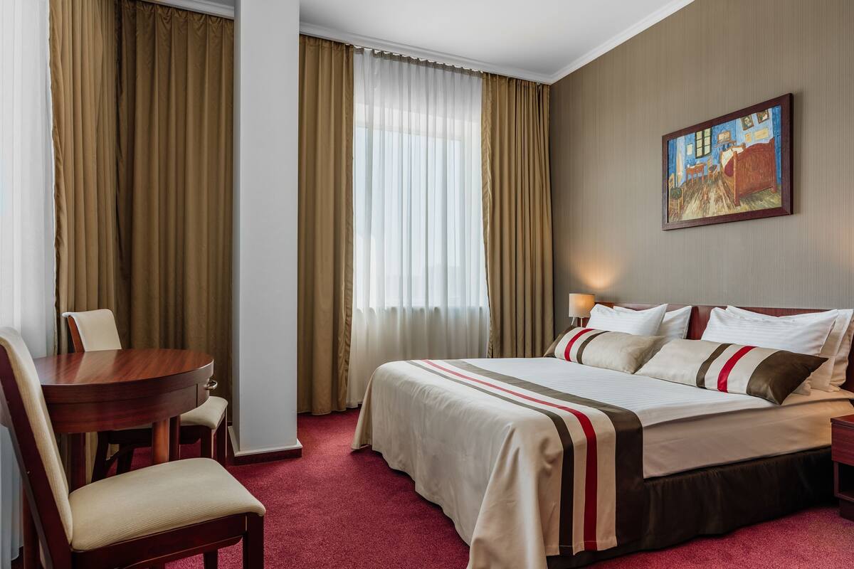 Une chambre d’hôtel avec un lit, un bureau, une chaise, un tableau au mur et une fenêtre avec des rideaux.