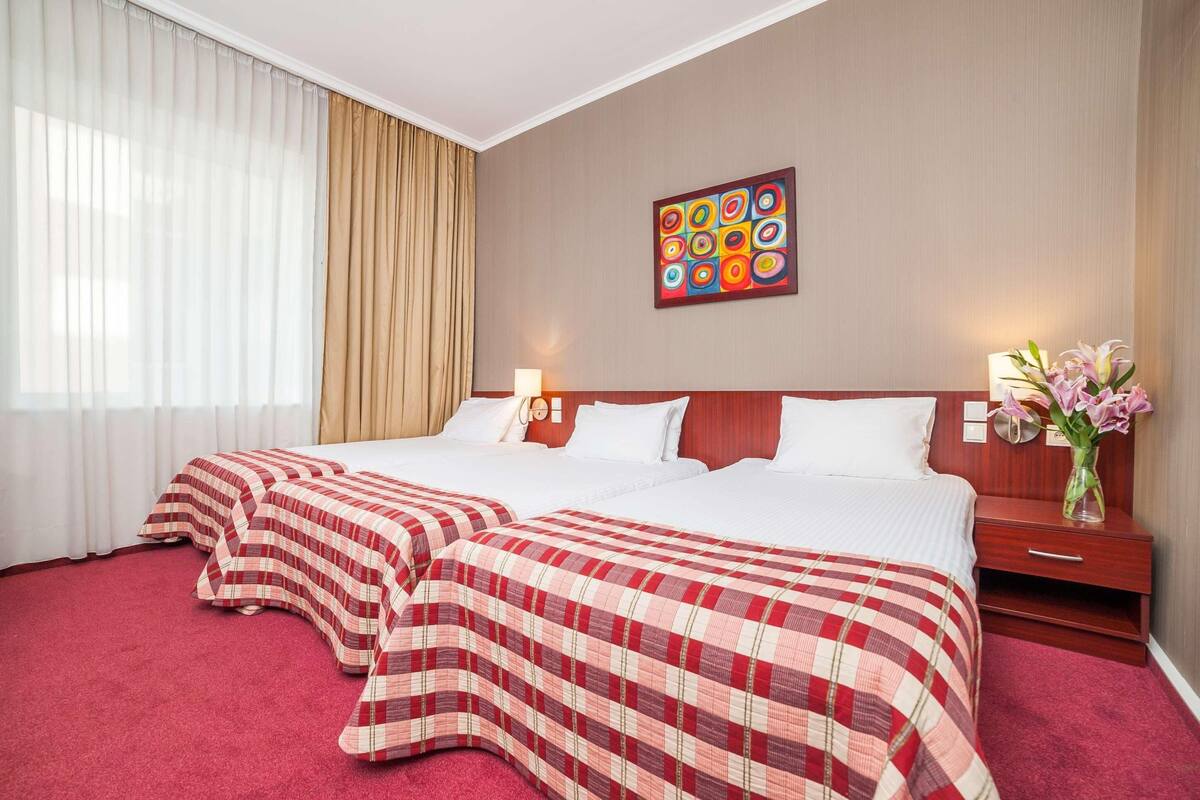 Une chambre d’hôtel avec deux lits, une grande fenêtre avec des rideaux, un tableau décoratif au mur, une table de chevet avec une lampe et un vase rempli de fleurs.