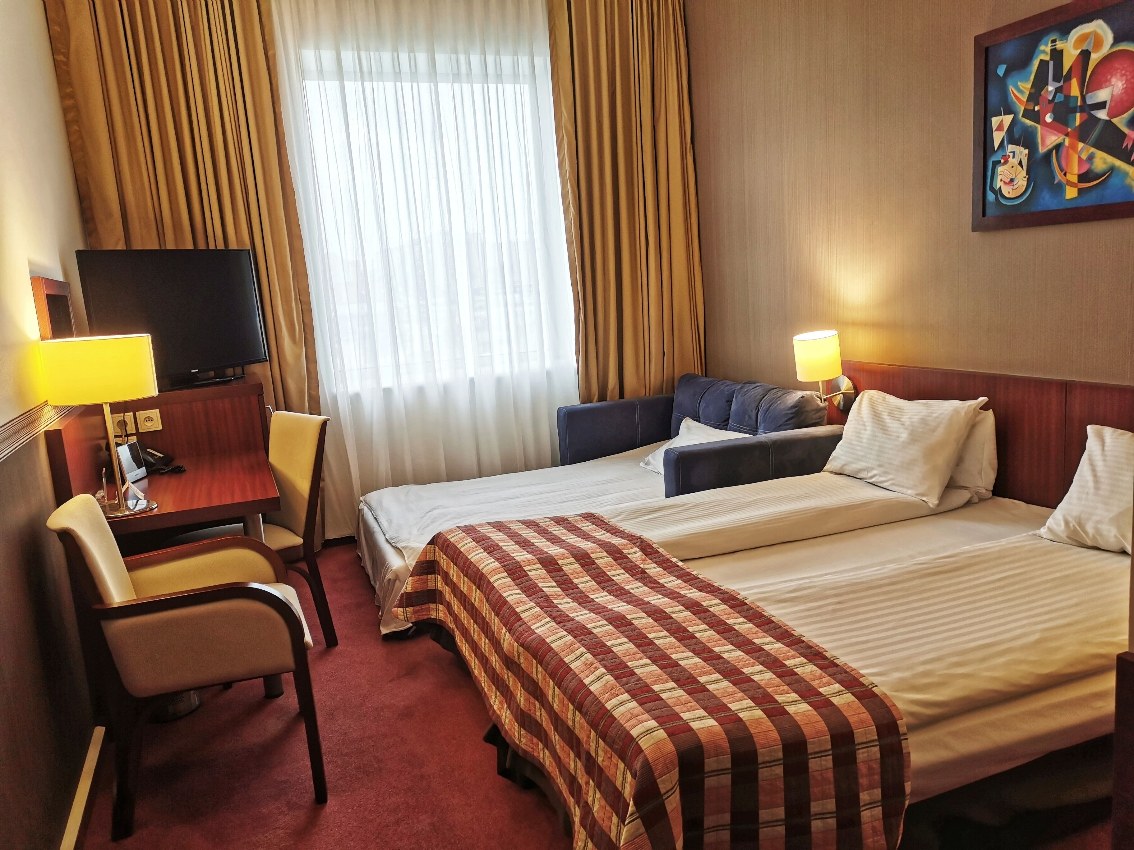 A hotel room with a bed, a desk with a lamp, a chair, a television, and a painting on the wall.