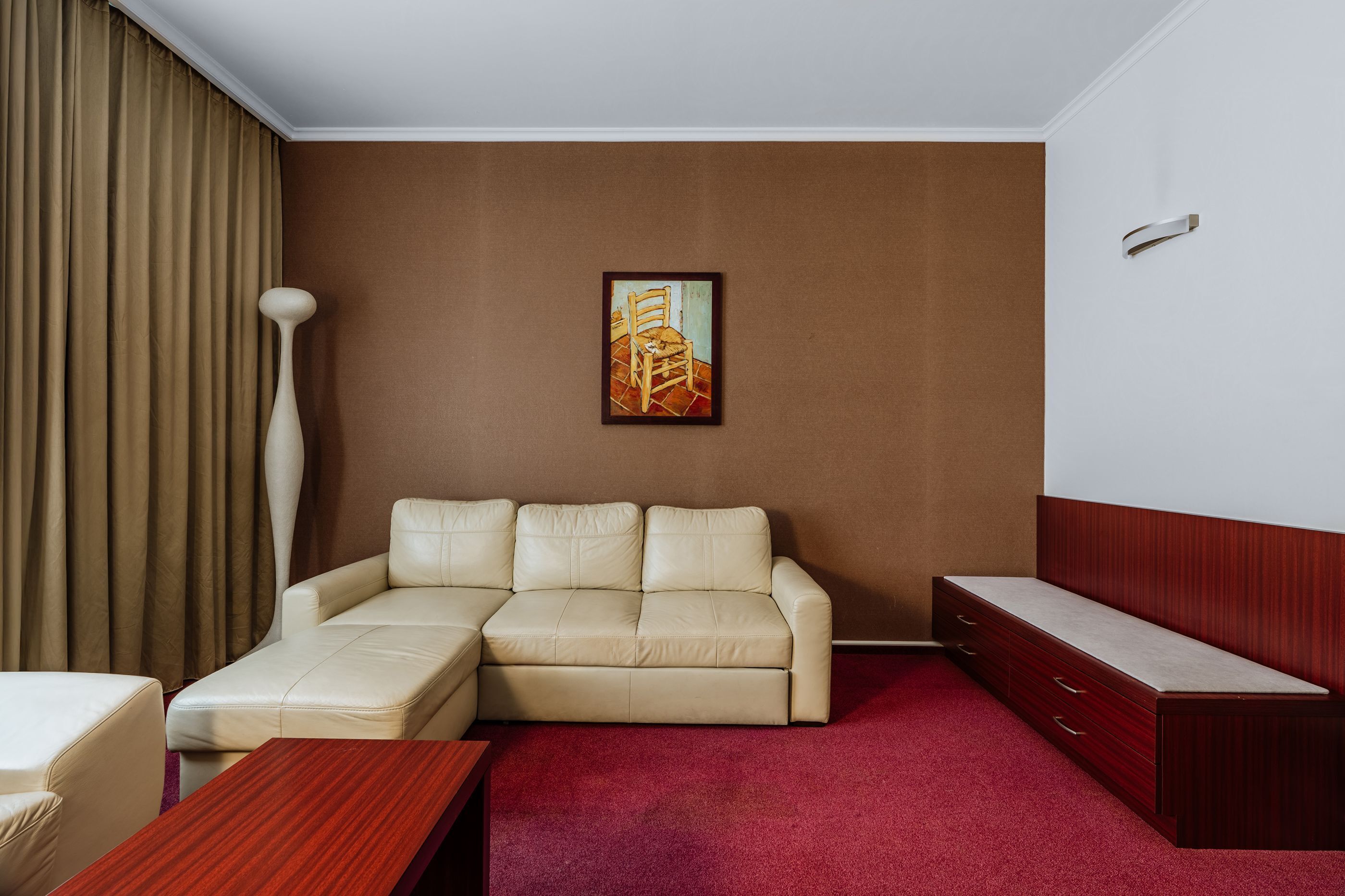 A hotel room with a beige sectional sofa, a wooden coffee table, a wooden side table, a red carpet, a painting on the wall, and a large floor lamp.