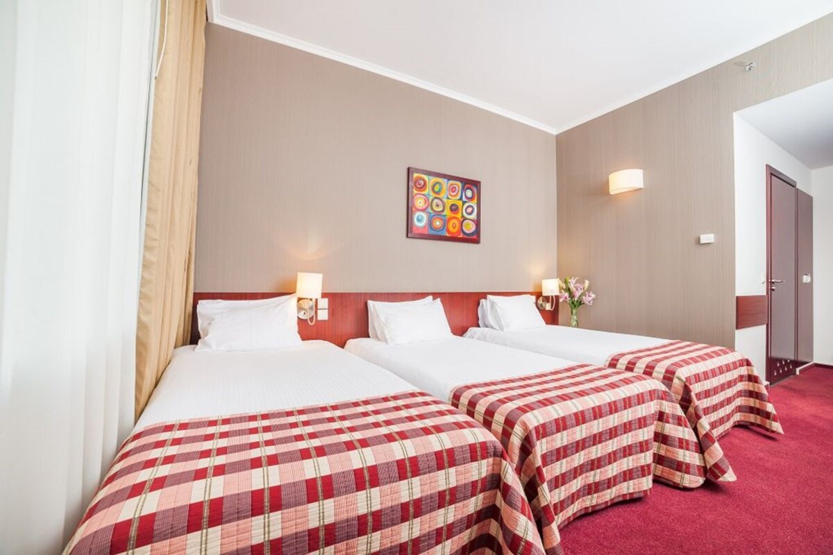 A hotel room with three beds, each with a red and white checkered bedspread. There is a painting on the wall above the beds and a vase with flowers on the nightstand.
