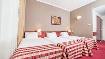 Une chambre d’hôtel avec trois lits, chacun recouvert d’une couvre-lit à carreaux rouges et blancs. Un tableau est accroché au mur au-dessus des lits, et un vase rempli de fleurs est posé sur la table de chevet.
