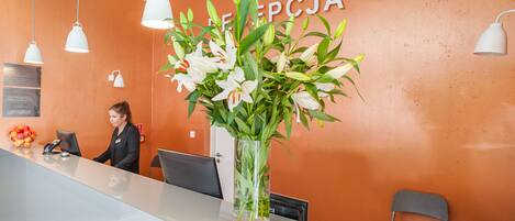 Una mesa de recepción con un jarrón grande de flores, una recepcionista y un cartel en la pared.