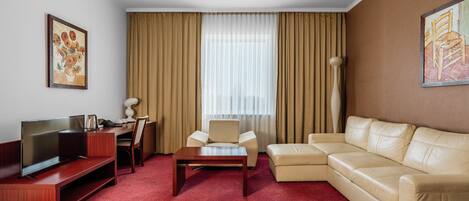 A hotel room with a TV, a sofa, a chair, a coffee table, and a large window with curtains.