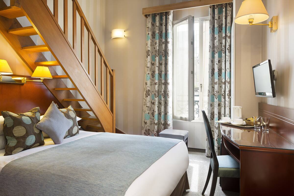 A hotel room with a bed, a desk, a chair, a television, a window with curtains, a staircase, and a wall-mounted lamp.