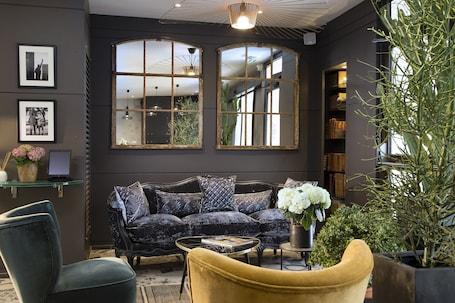 A modern living room with a dark-colored sofa, a yellow armchair, and a glass coffee table. There are two large windows with decorative frames, a bookshelf, and a potted plant.