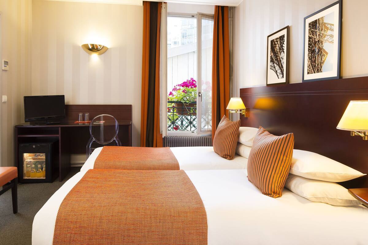 A hotel room with a bed, two bedside lamps, a television, a desk with a computer, and a window with curtains.