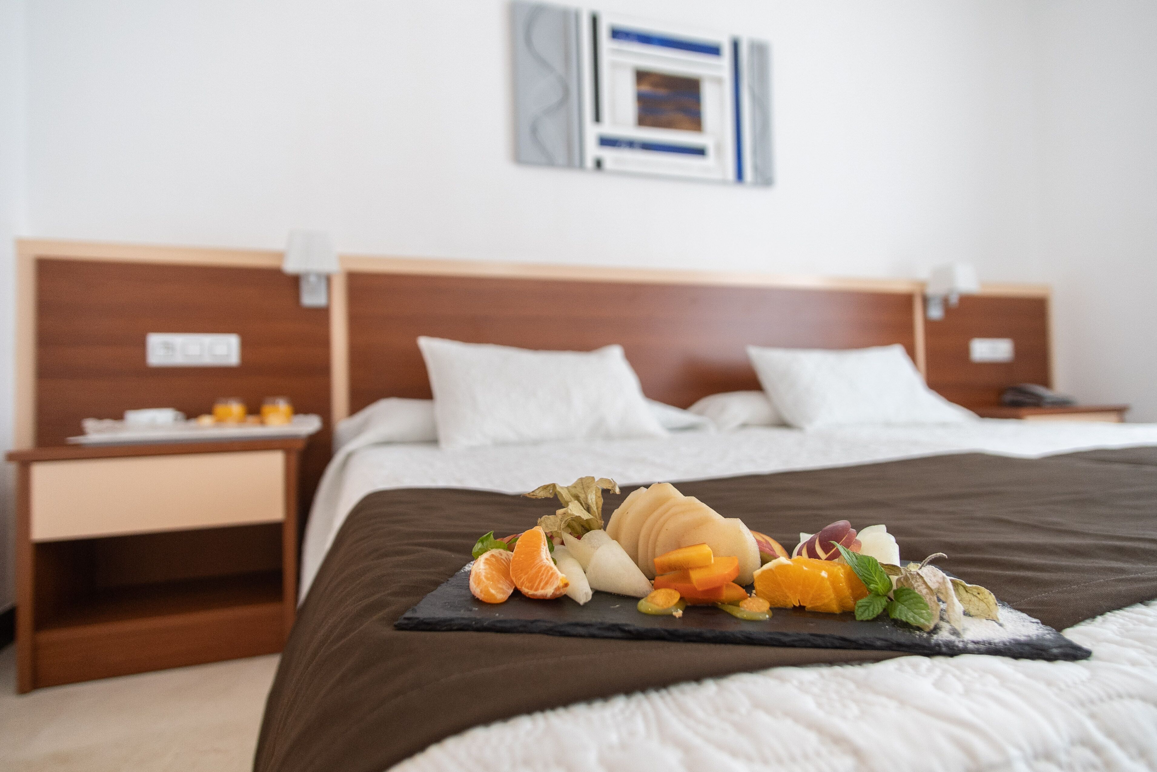 Una cama bien hecha con una bandeja de frutas sobre el edredón, un cabecero de madera y una mesita de noche.