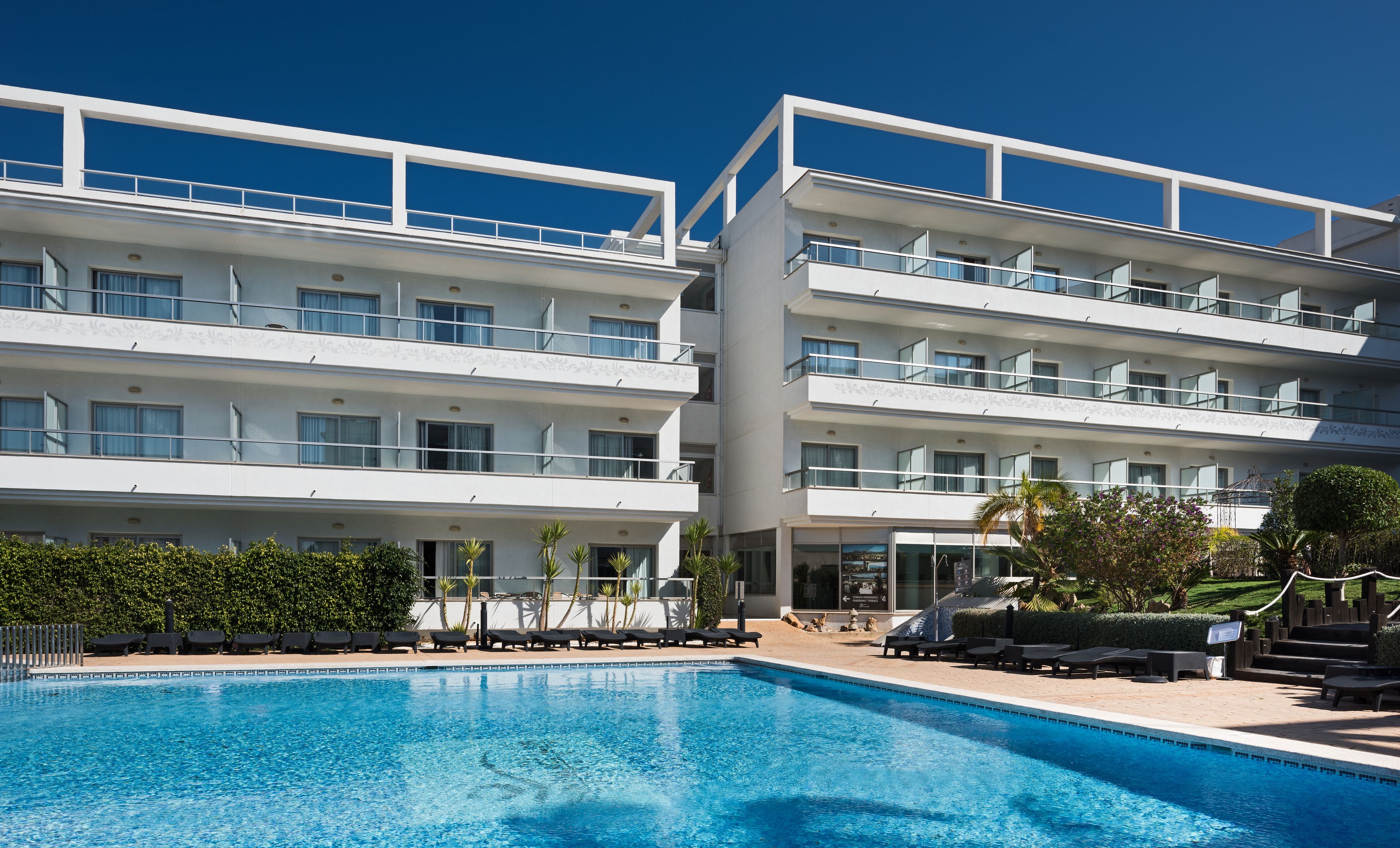 Un hotel moderno con zona de piscina, varios balcones y un jardín bien cuidado.