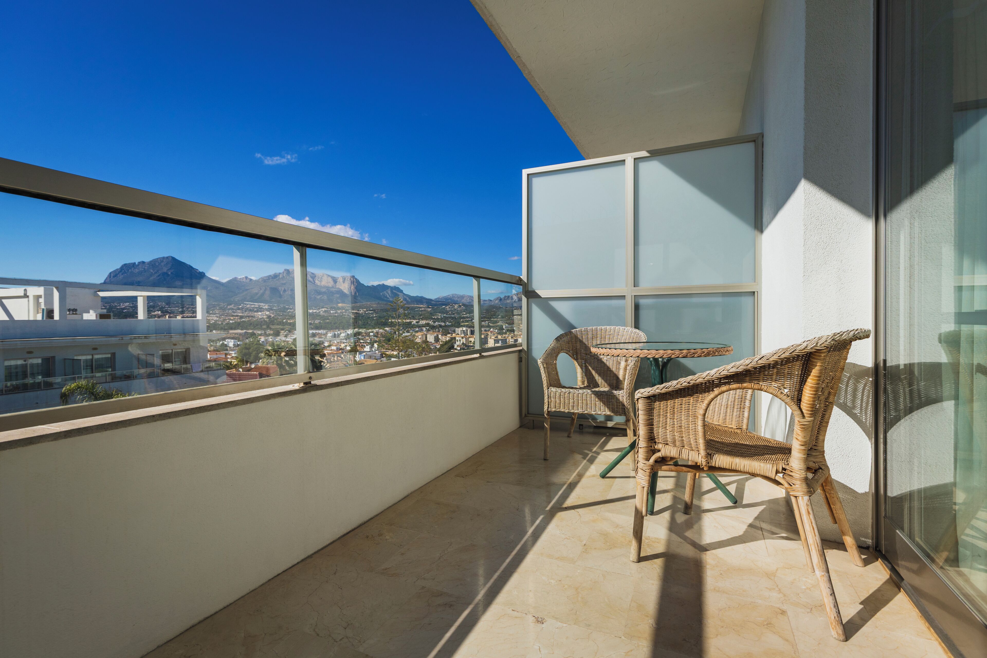 Un balcón con sillas de mimbre, una mesa y vistas a montañas y edificios.