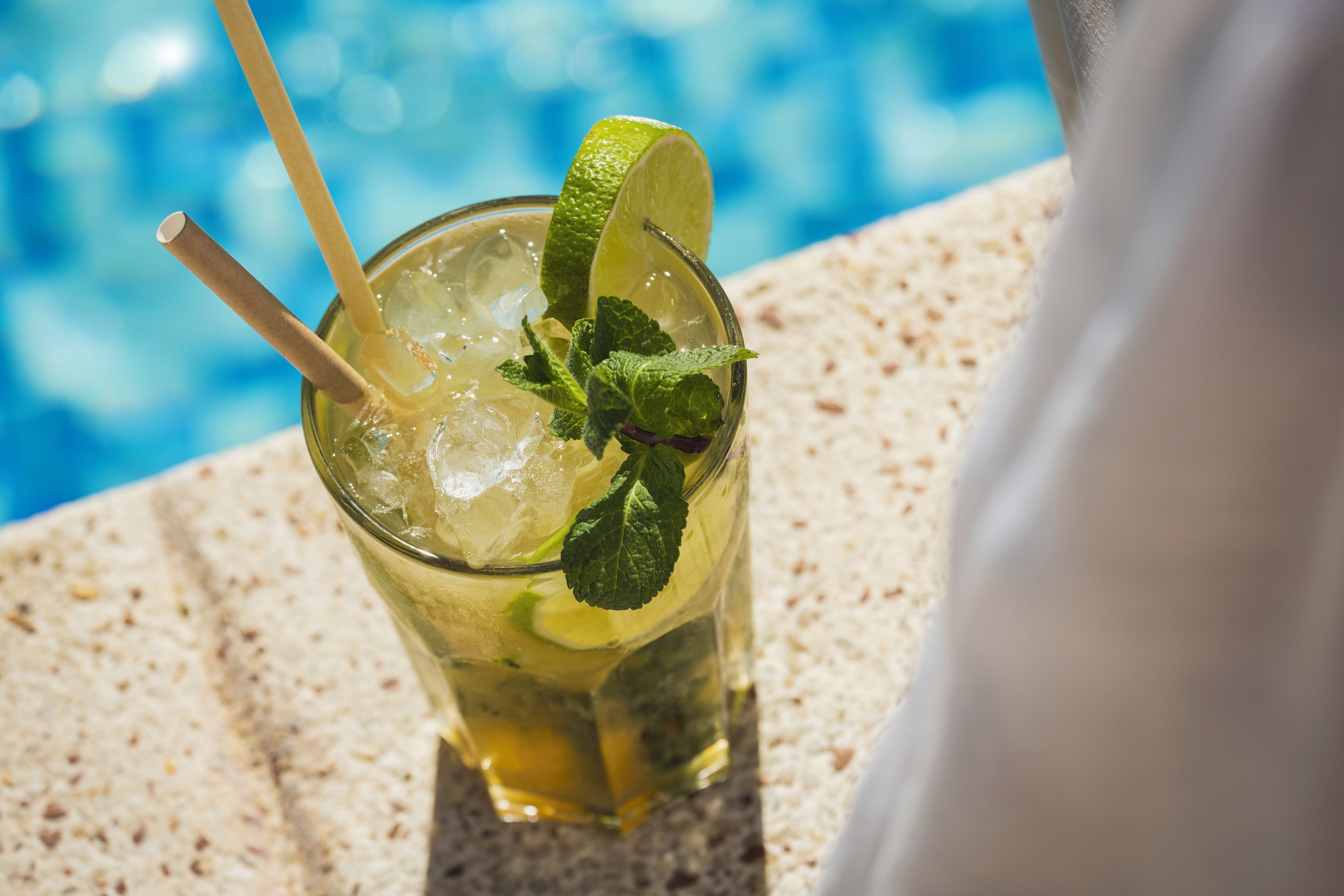 A glass of a beverage with a lime wedge and mint garnish, placed on a textured surface near a pool.