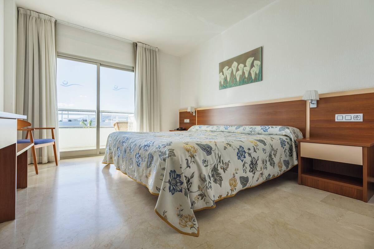 A hotel room with a bed, a desk, a chair, a window with curtains, and a painting on the wall.