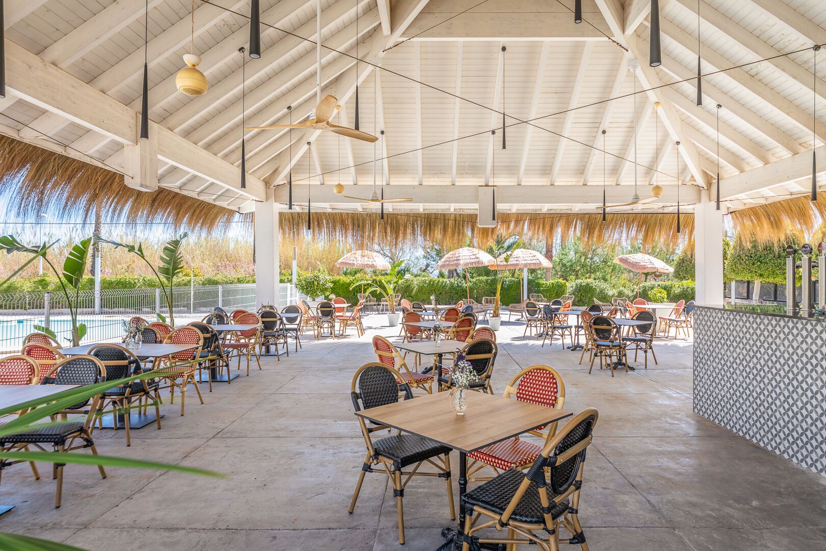 An outdoor dining area with tables and chairs, a pool in the background, and a covered ceiling with hanging lights.