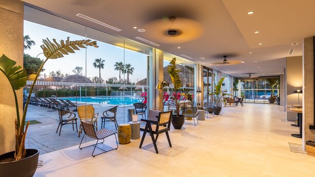 Un moderno vestíbulo de hotel con vista a la piscina, asientos interiores y plantas en macetas.