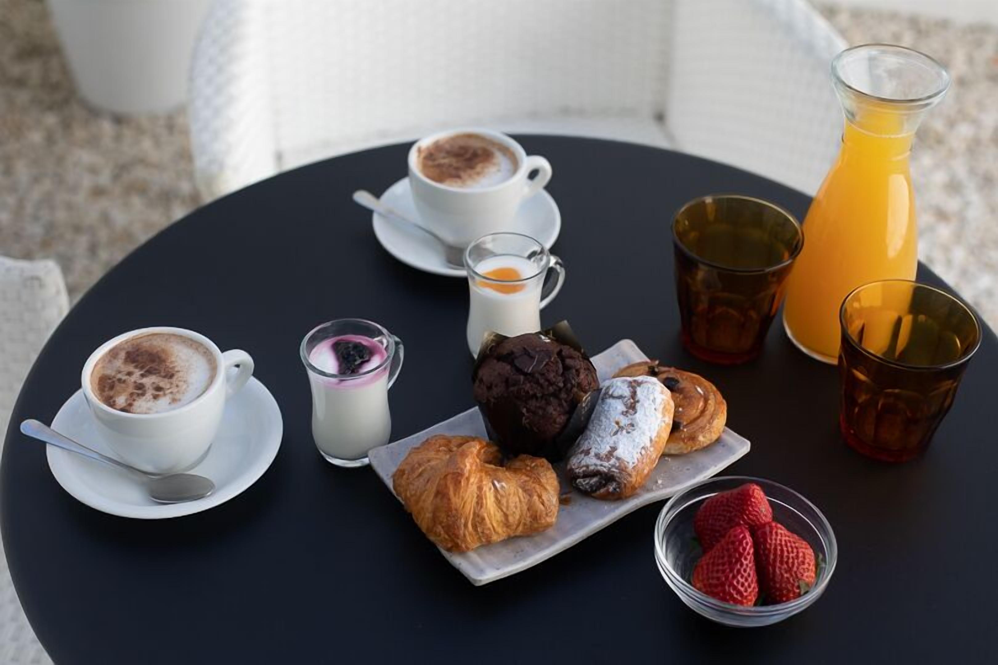 Une table avec des tasses à café, des pâtisseries, un verre de lait, un verre de jus d’orange et un bol de fraises.