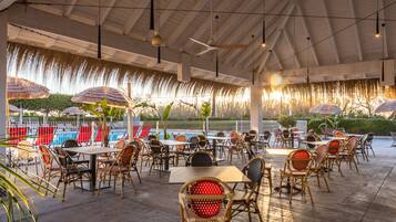 An outdoor dining area with tables and chairs, a pool in the background, and a ceiling fan.