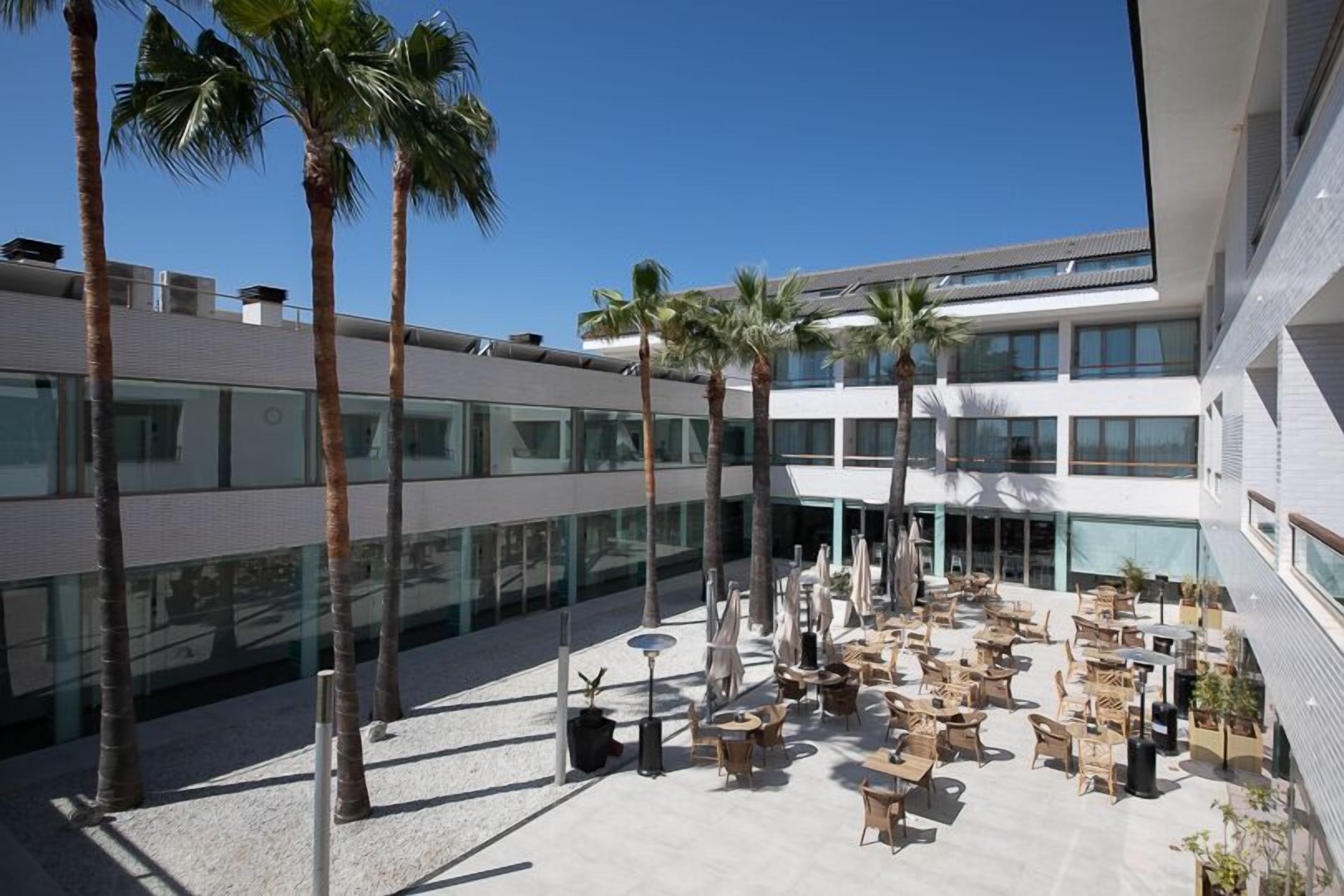 Un bâtiment moderne doté d’une cour intérieure aménagée avec des espaces de détente en plein air, des palmiers et de grandes fenêtres.