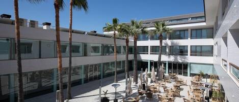 Un edificio moderno con un patio que cuenta con mesas al aire libre, palmeras y ventanas amplias.