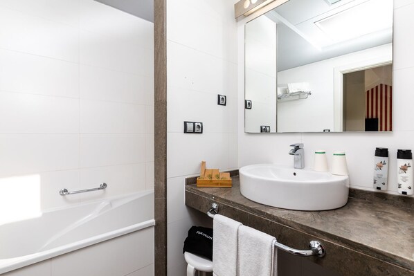 A modern bathroom with a white bathtub, a large mirror, and a vanity with a sink and toiletries. - Hotel Barú (Sagunto)