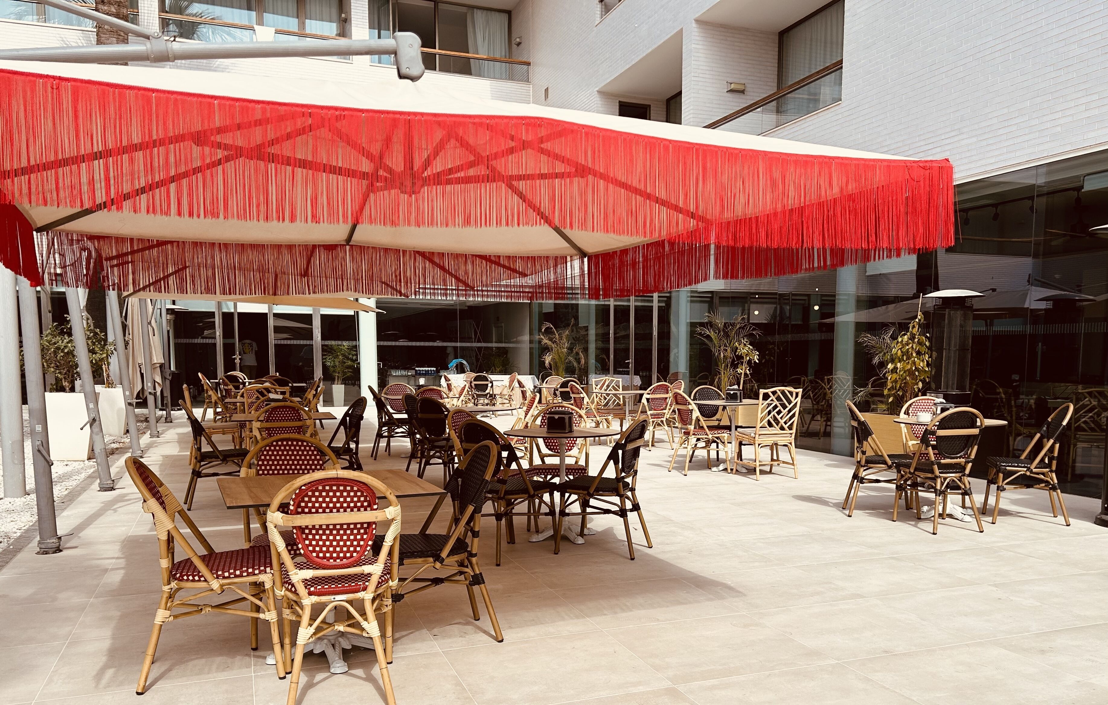 Un espace extérieur aménagé avec des chaises et des tables en osier, protégé par un auvent rouge, et un bâtiment en verre en arrière-plan.