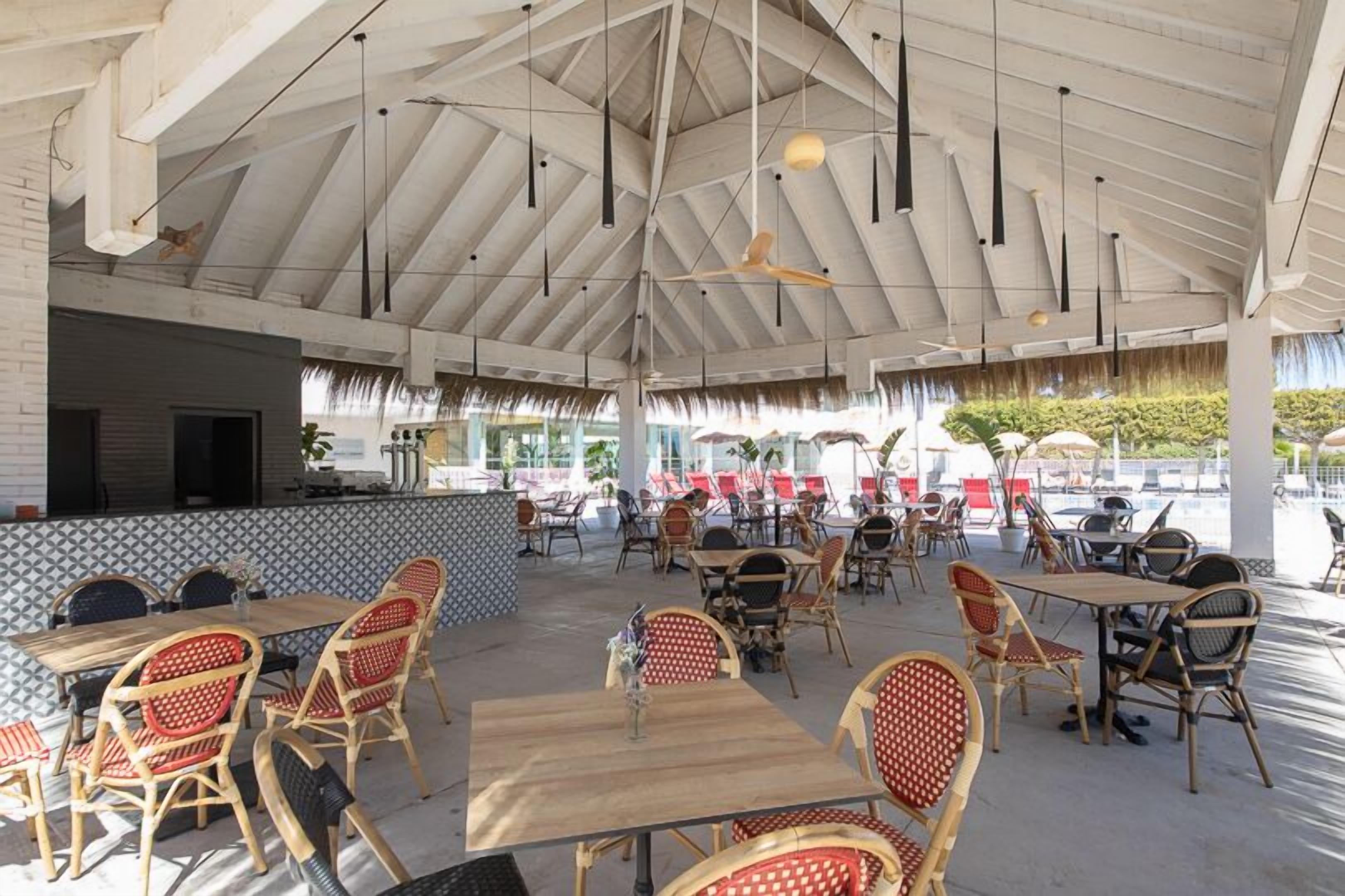 An outdoor dining area with wooden tables and wicker chairs, a high ceiling with exposed beams, and a view of the pool area in the background.