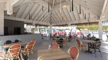An outdoor dining area with wooden tables and wicker chairs, a high ceiling with exposed beams, and a view of the pool area in the background.