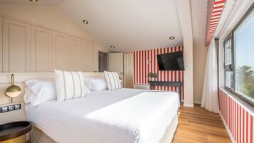 Habitación de hotel con una cama grande, una mesita de noche con lámpara, un televisor montado en la pared y una ventana con cortinas.